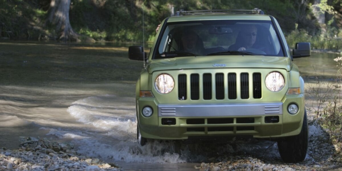 Khaki Jeep patriot driving across a tiny river with the headlights on